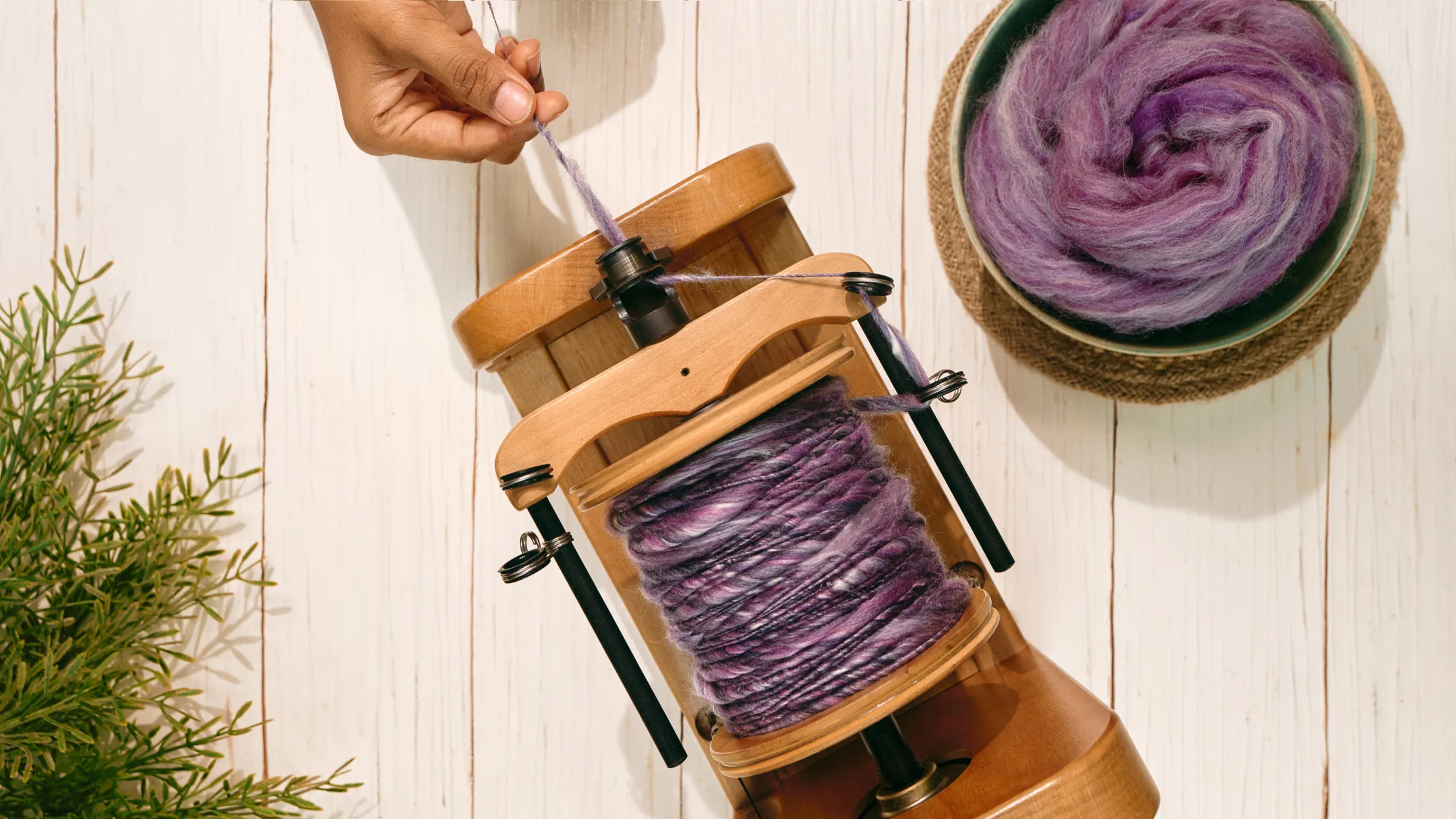 A wooden spinning wheel bobbin filled with purple handspun yarn beside a bowl of roving
