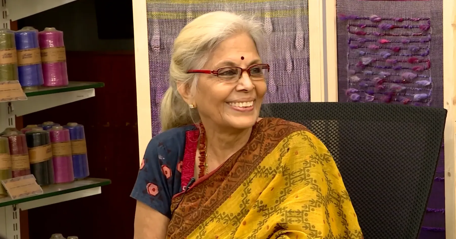 Mrs. Padma Srinath smiling in front of handwoven textiles in the studio