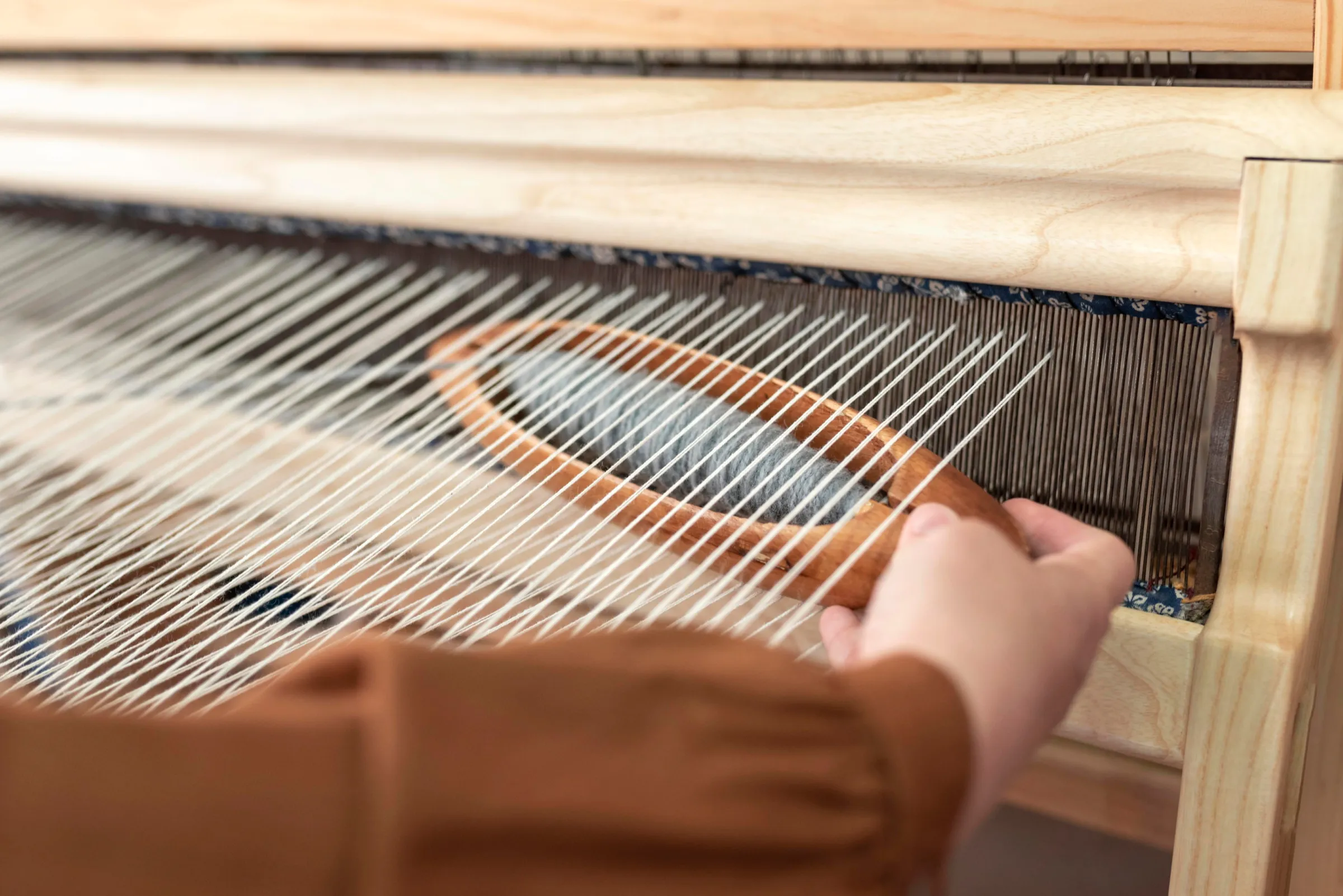 A hand passes a wooden shuttle through the white warp of a table loom