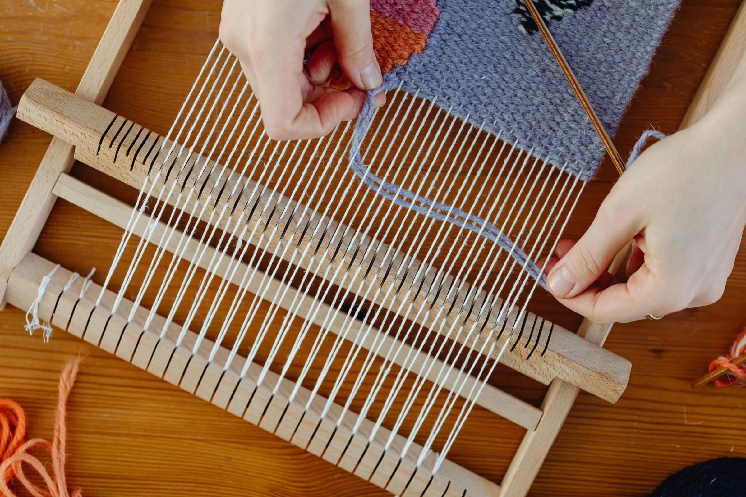 Hands weaving yarn through the warp of a small wooden frame loom
