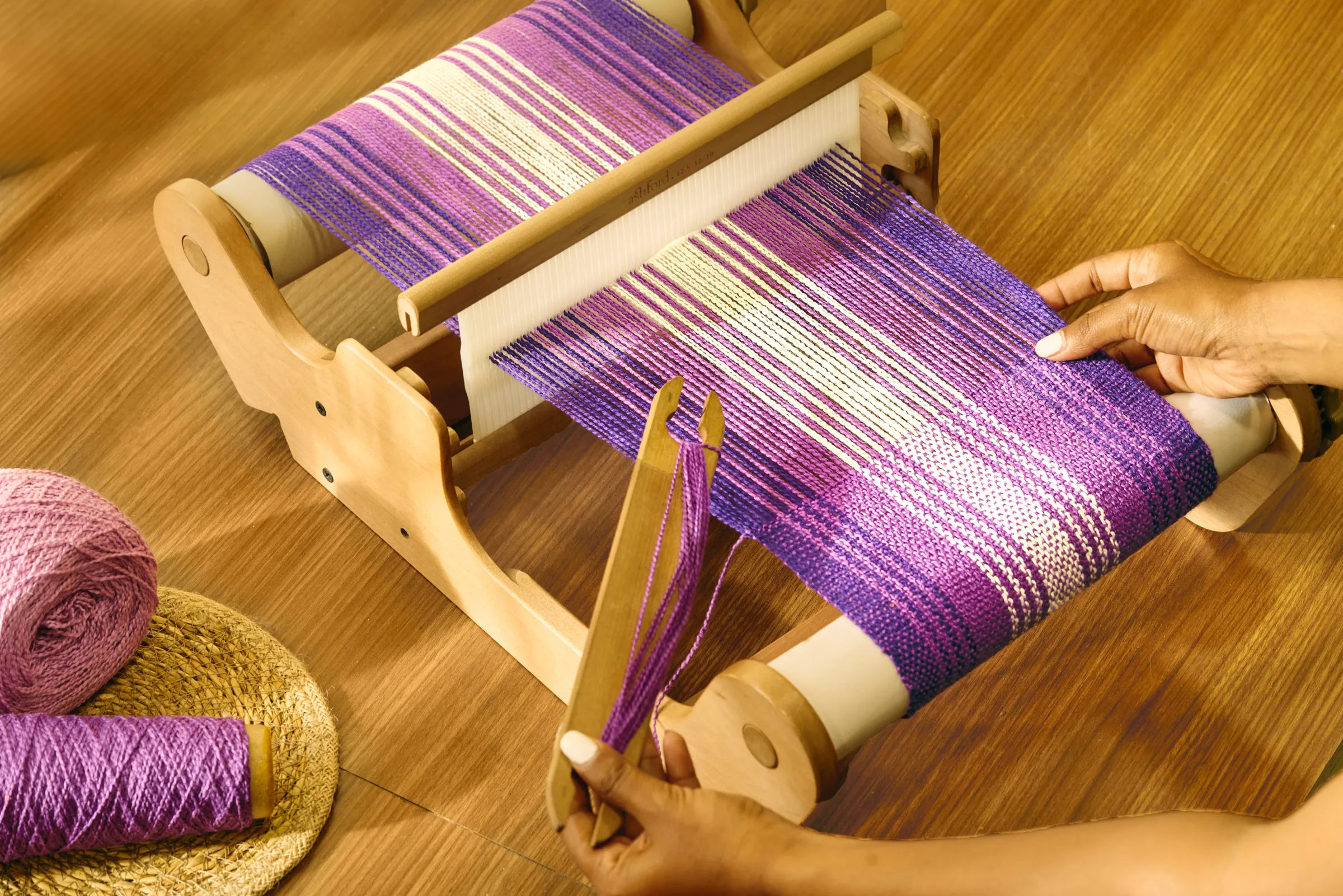 Hands weaving purple and white yarn on a rigid heddle loom