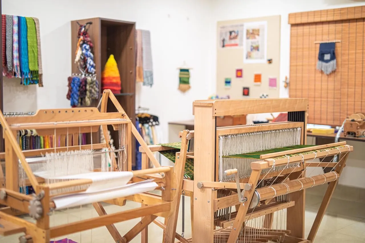 Wooden table and floor looms in the studio in front of shelves of yarn