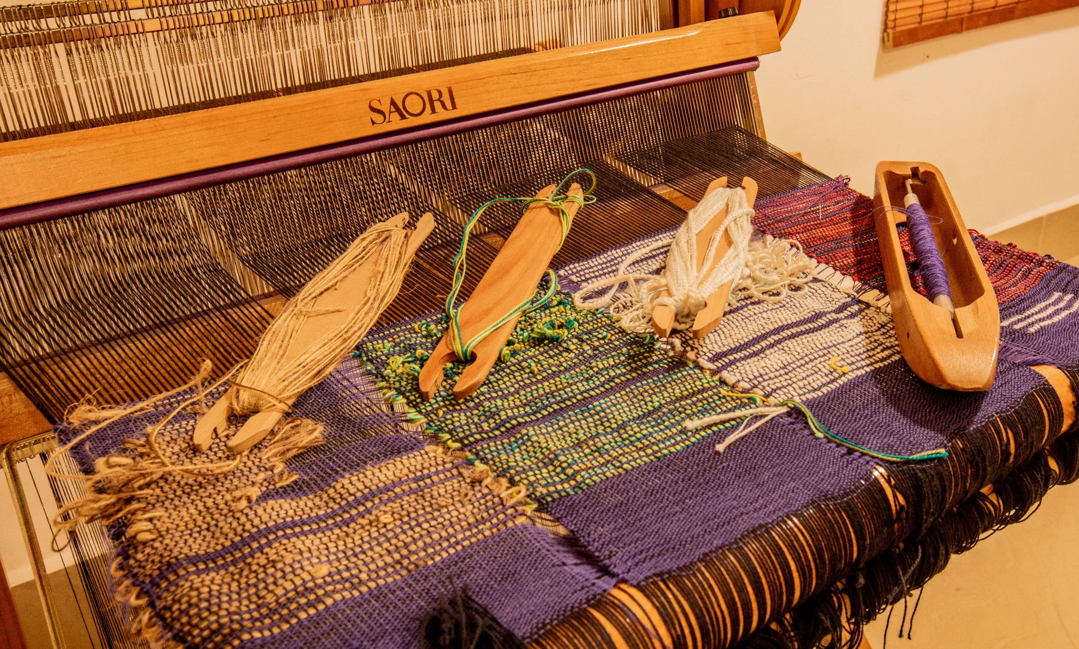 Wooden SAORI shuttles wound with yarn resting on purple handwoven cloth on the loom