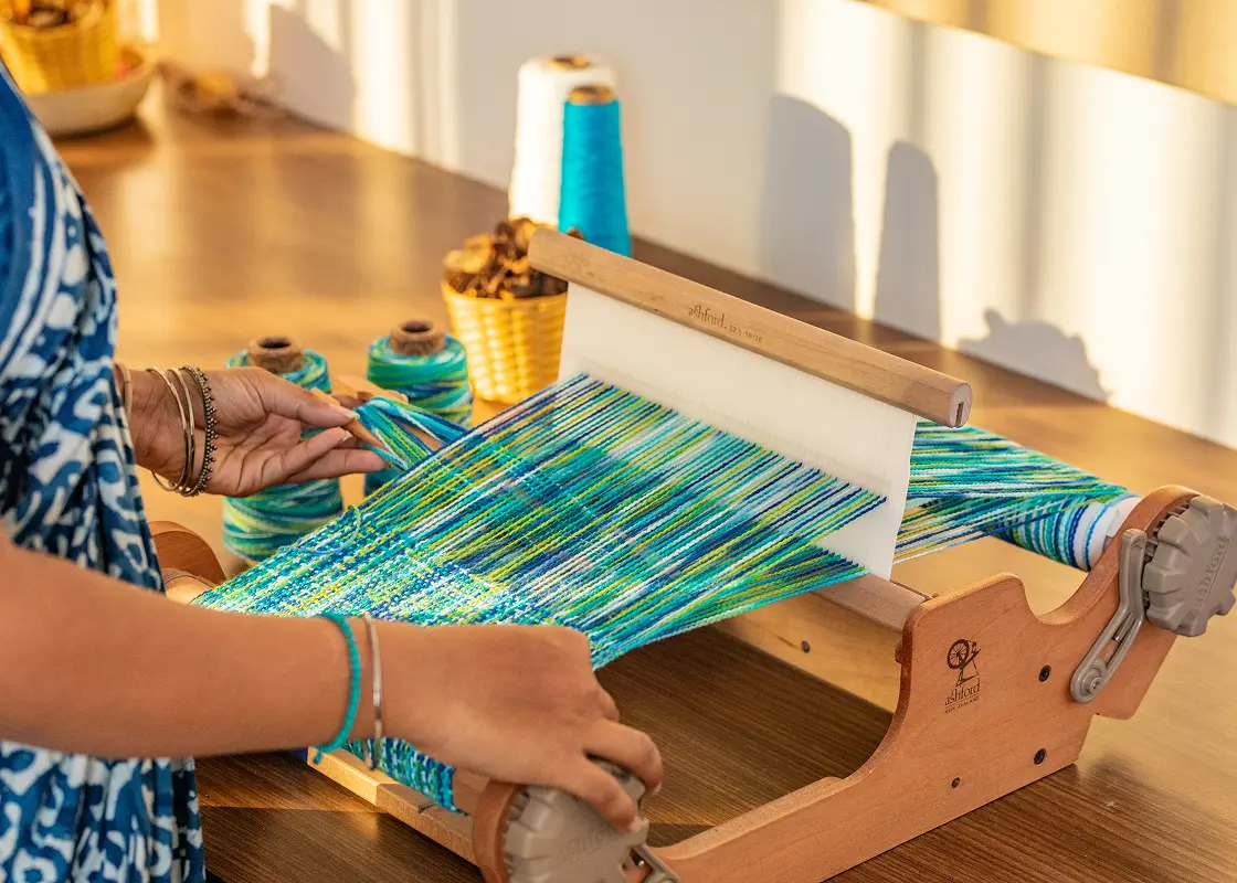 A weaver at a rigid heddle loom with a teal and green warp