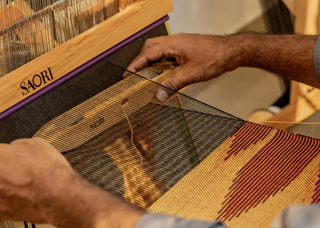 Hands beating the weft on a SAORI loom with a red and ochre cloth