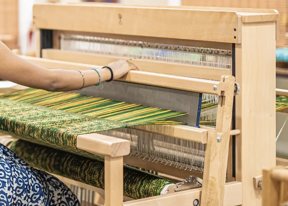A weaver at a wooden floor loom with a green and gold warp