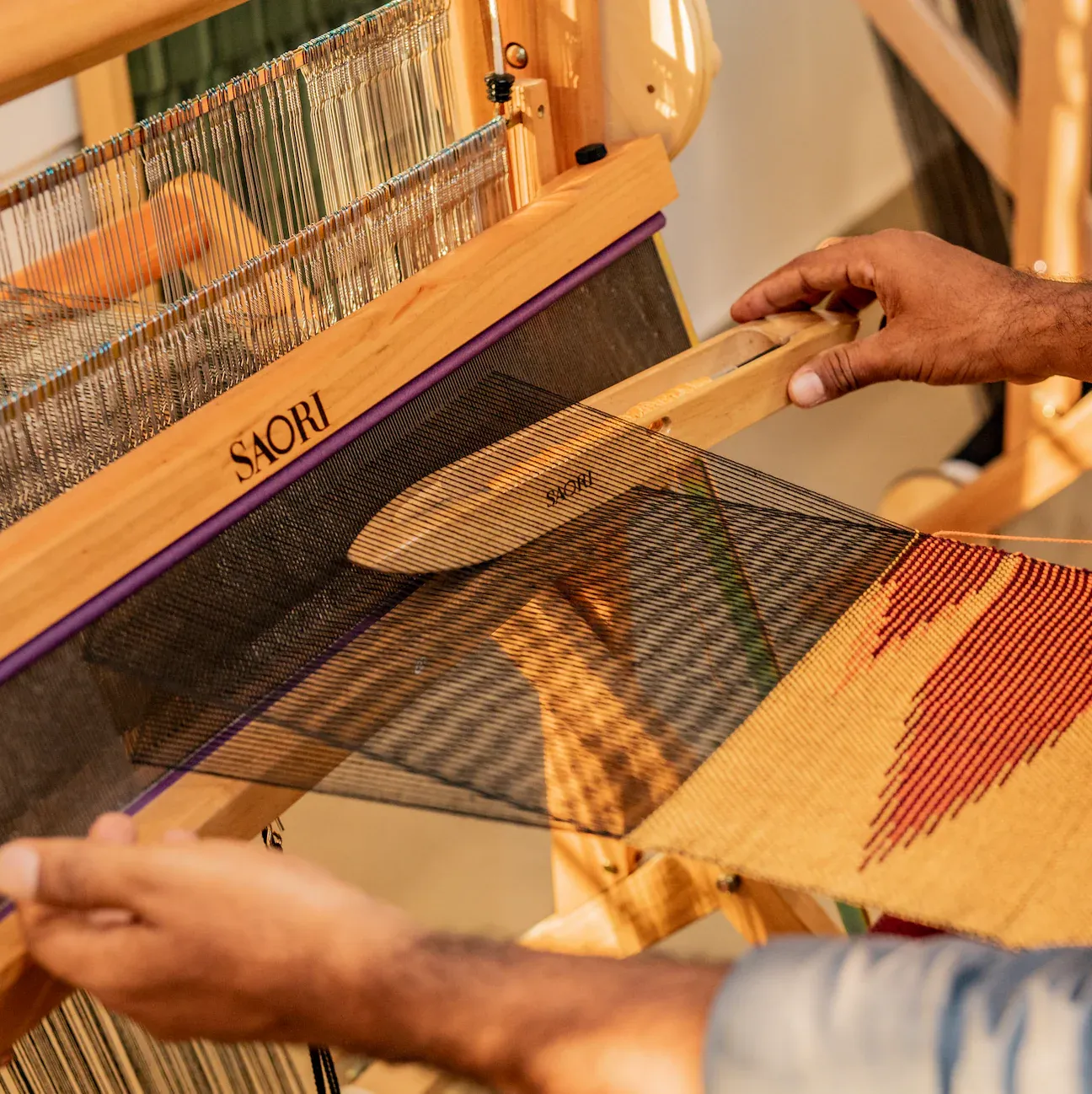 Hands weaving an orange and black cloth on a SAORI loom