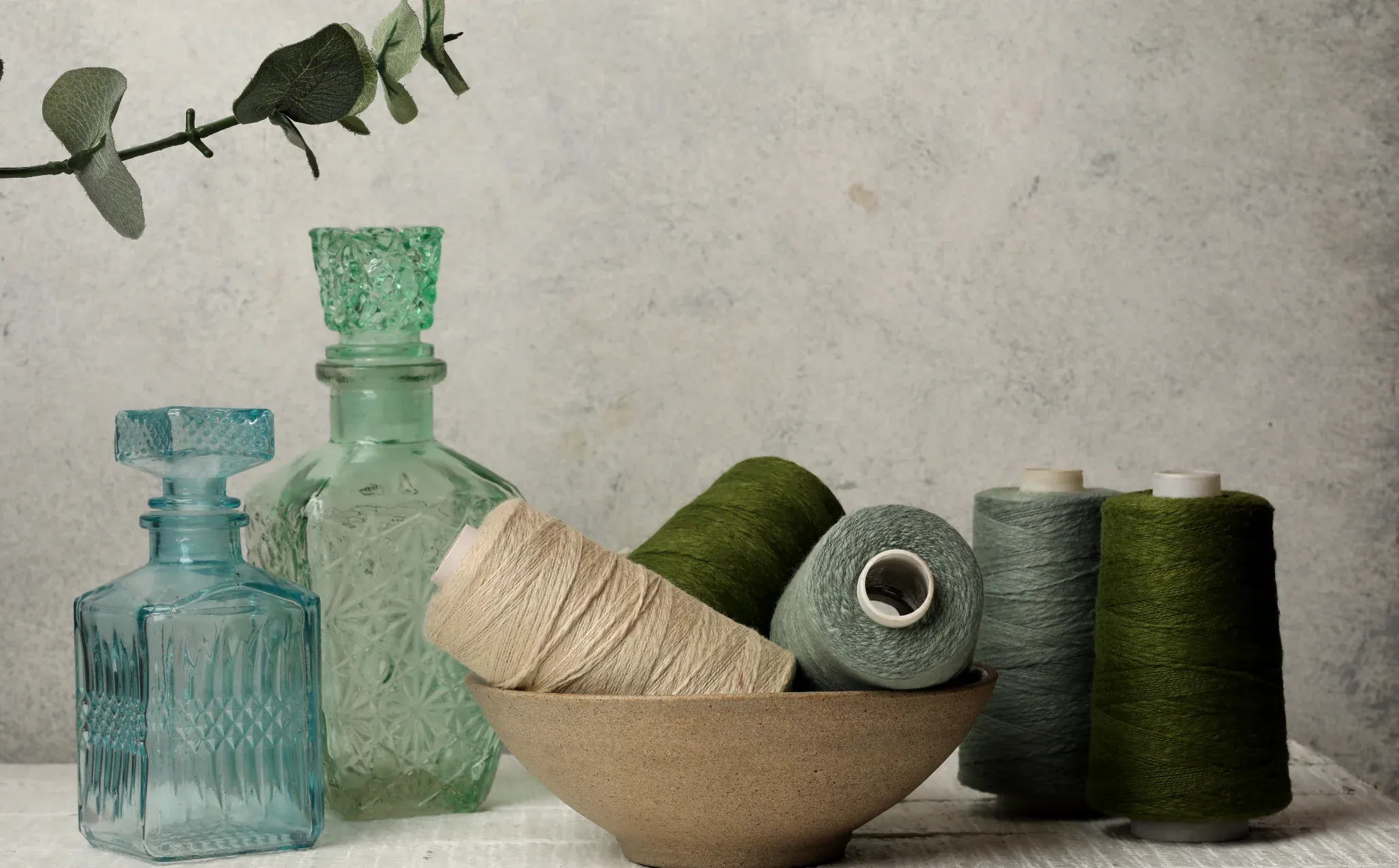 Cones of natural and green linen yarn in a stoneware bowl beside glass bottles