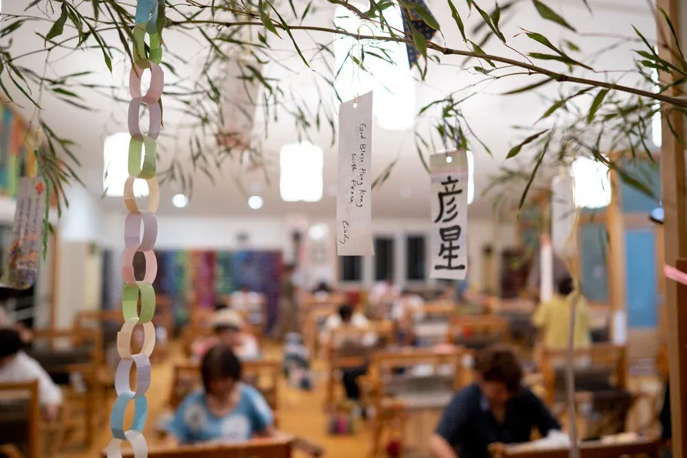 People weaving at rows of SAORI looms in a bright studio hung with paper decorations