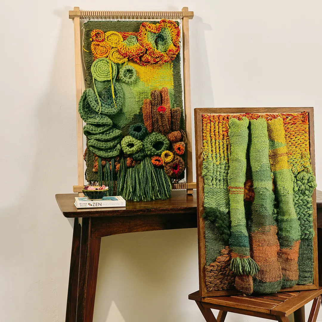 Two textured handwoven wall hangings in greens and oranges on a wooden side table