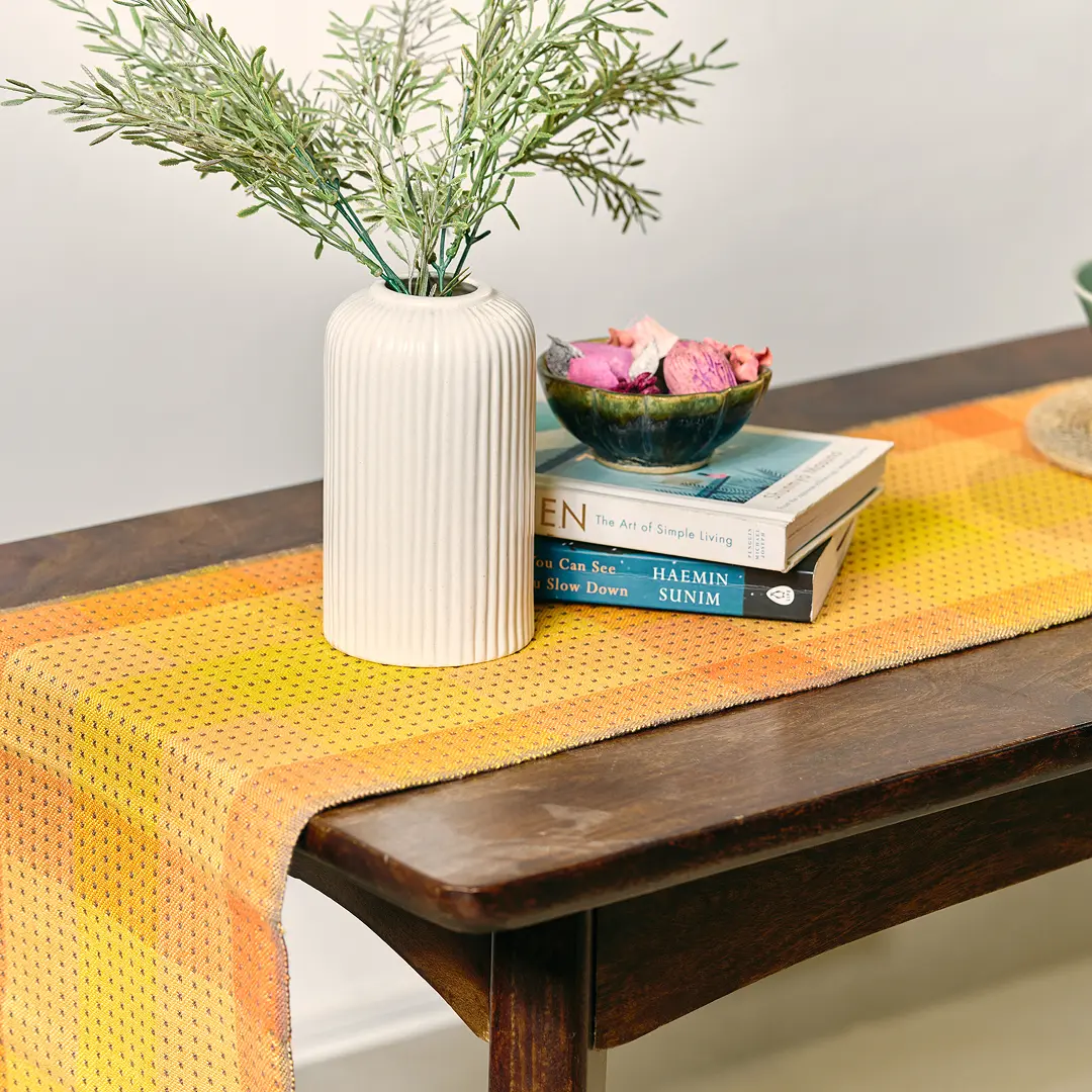 A handwoven orange and yellow table runner on a wooden console with a vase and books