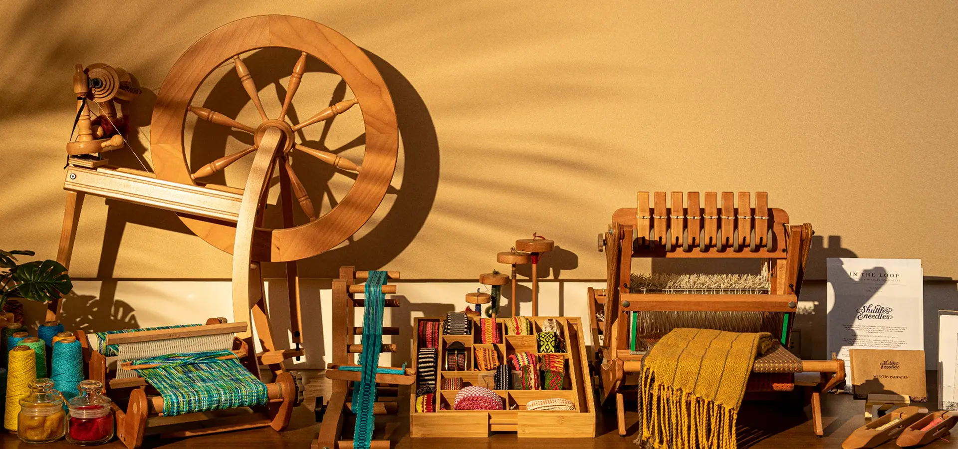 Spinning wheels, table looms, an inkle loom and a tray of woven bands in warm evening light