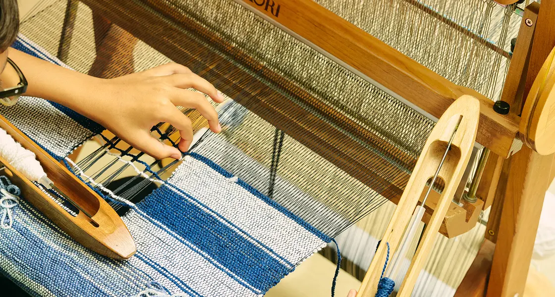 Hands passing a boat shuttle through a blue and white weave on a SAORI loom