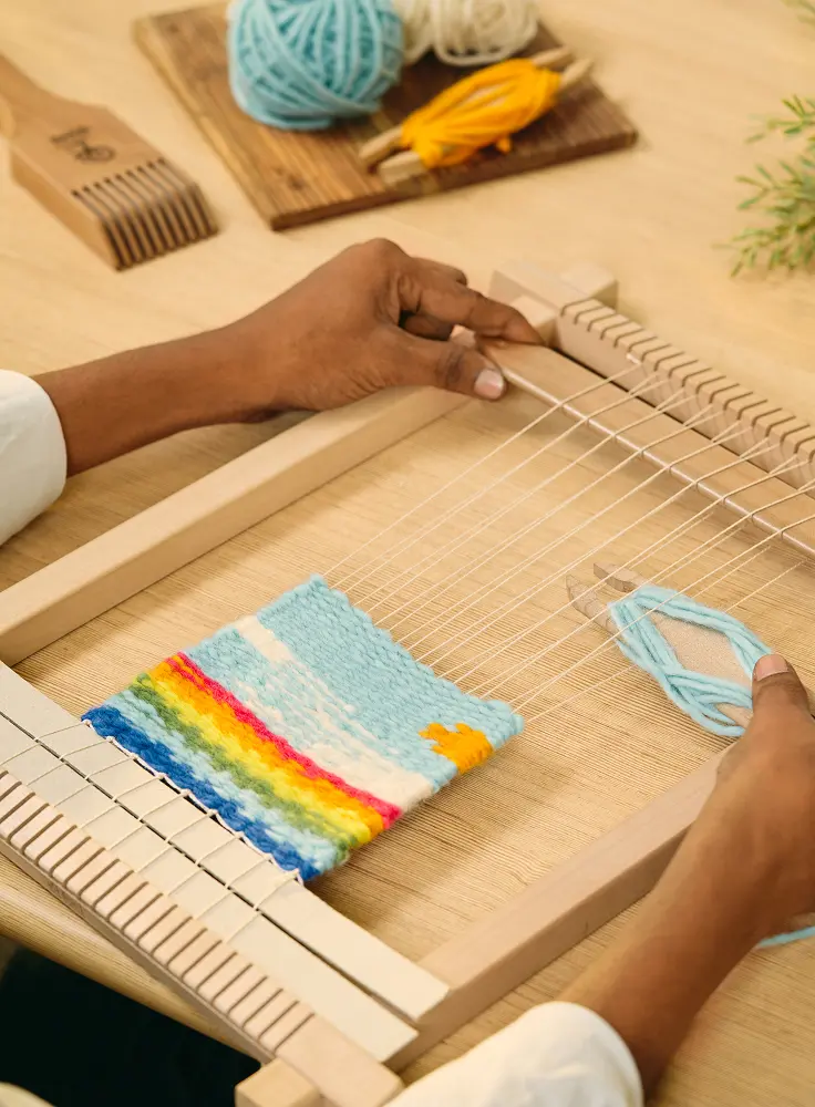 Hands weaving a rainbow-striped sample on a small wooden frame loom