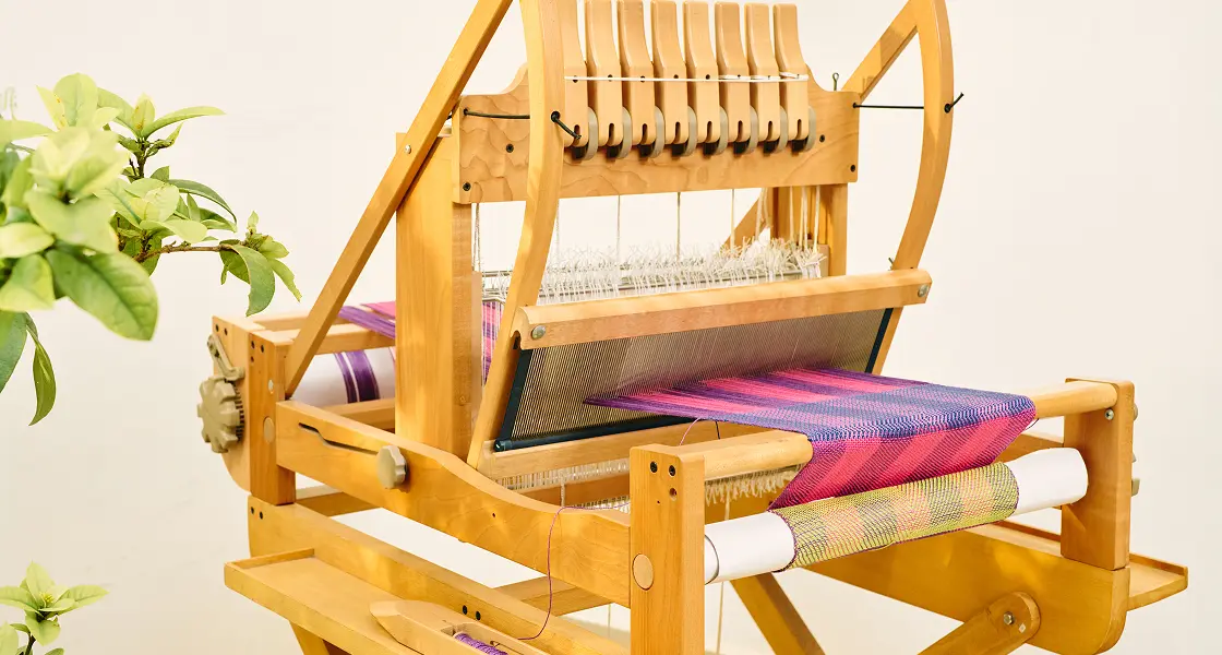 A multi-shaft table loom weaving a pink and purple cloth beside a potted plant