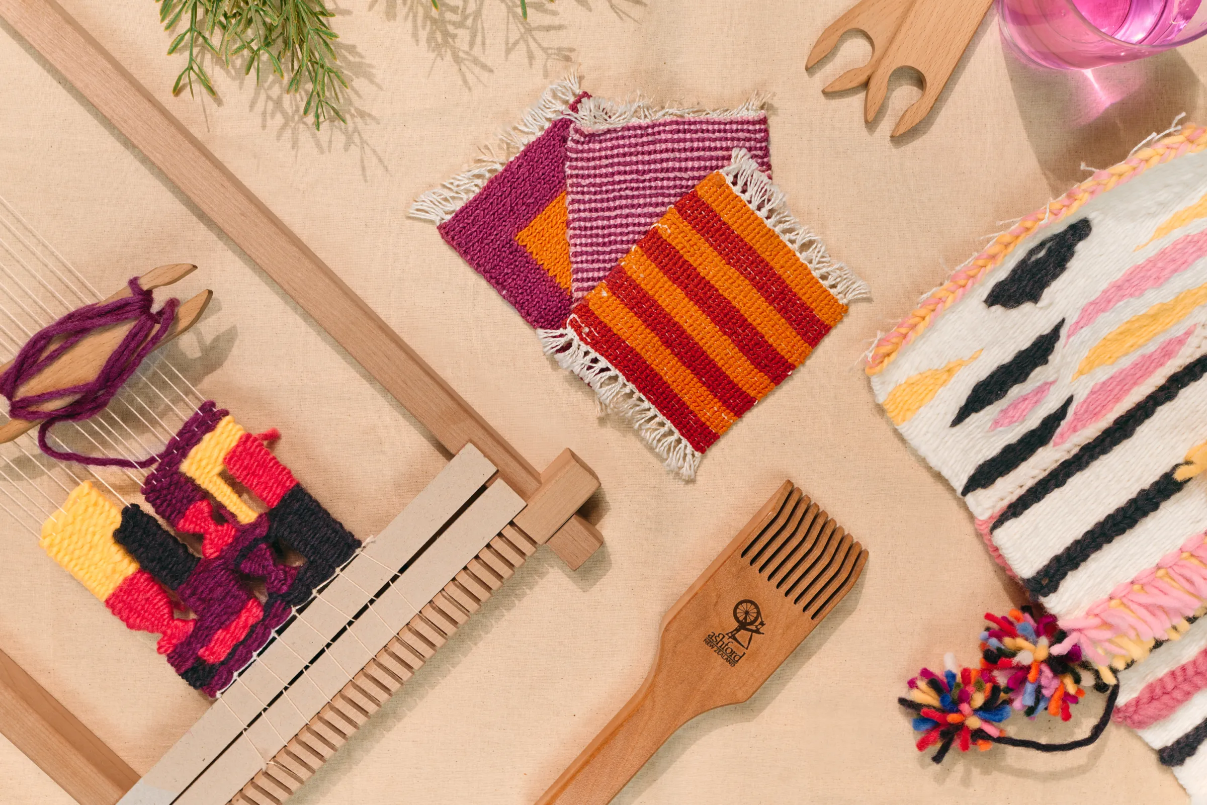 Weaving tools laid out on a table: a frame loom with a colourful woven piece, a wooden beater and striped weavings