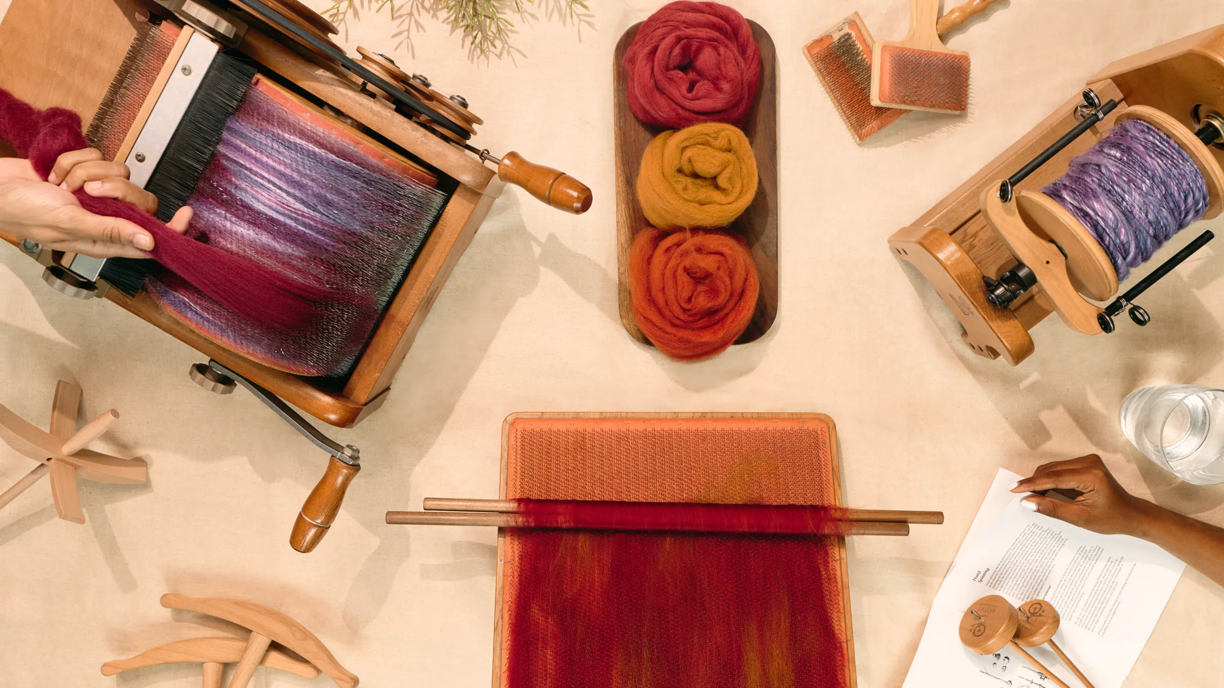 A drum carder, a spinning wheel and rolls of dyed wool roving on a table