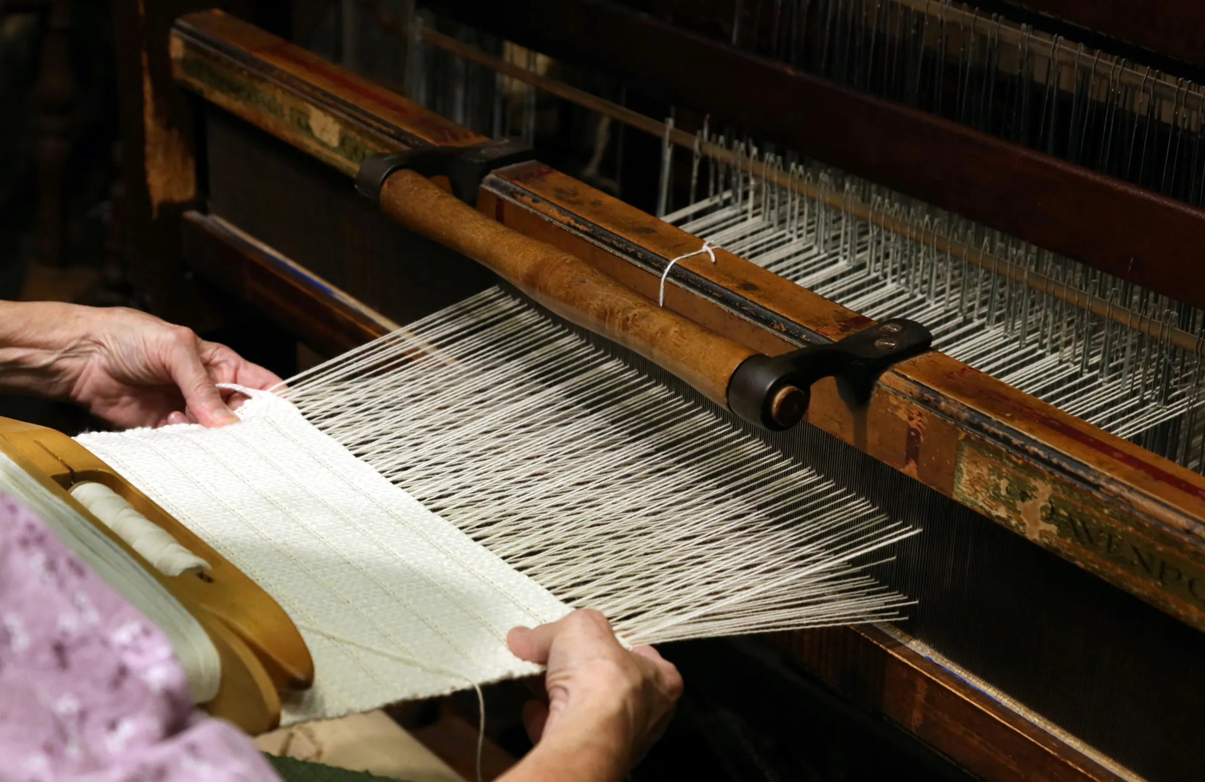 Hands weaving white cloth on a large wooden floor loom
