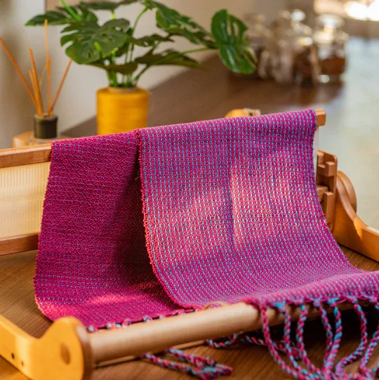 A magenta and blue handwoven scarf draped over a wooden loom