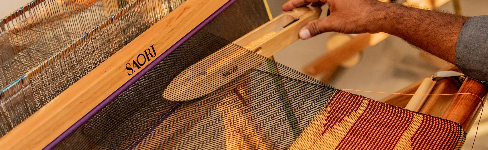 A weaver passes a shuttle through the warp of a SAORI loom