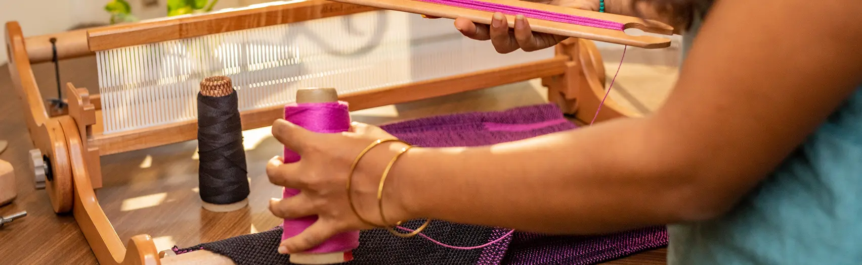 A student holds a cone of magenta yarn beside a rigid heddle loom