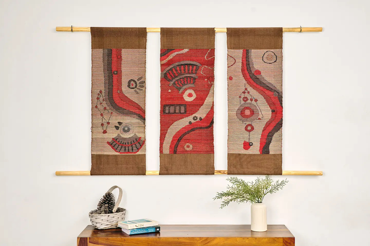 Three handwoven wall hangings in red, beige and brown above a wooden side table